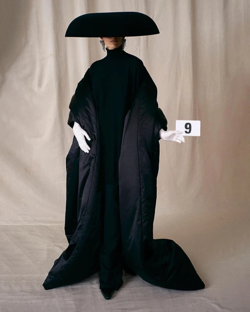 Fashionable adult poses in oversized black coat, turtleneck, and wide-brimmed hat in front of a neutral-toned background with a number nine.