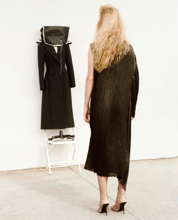 A woman with long curly hair wearing a dark dress and fishnet heels stands facing a black coat displayed on a white chair.