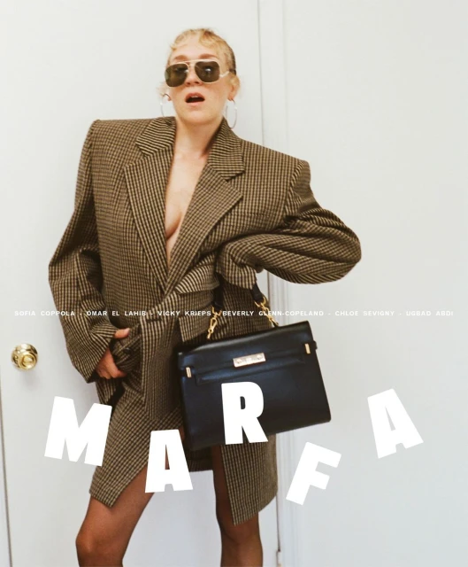 Model poses in an oversized brown houndstooth blazer dress and black tights, holding a black purse against a white wall background.