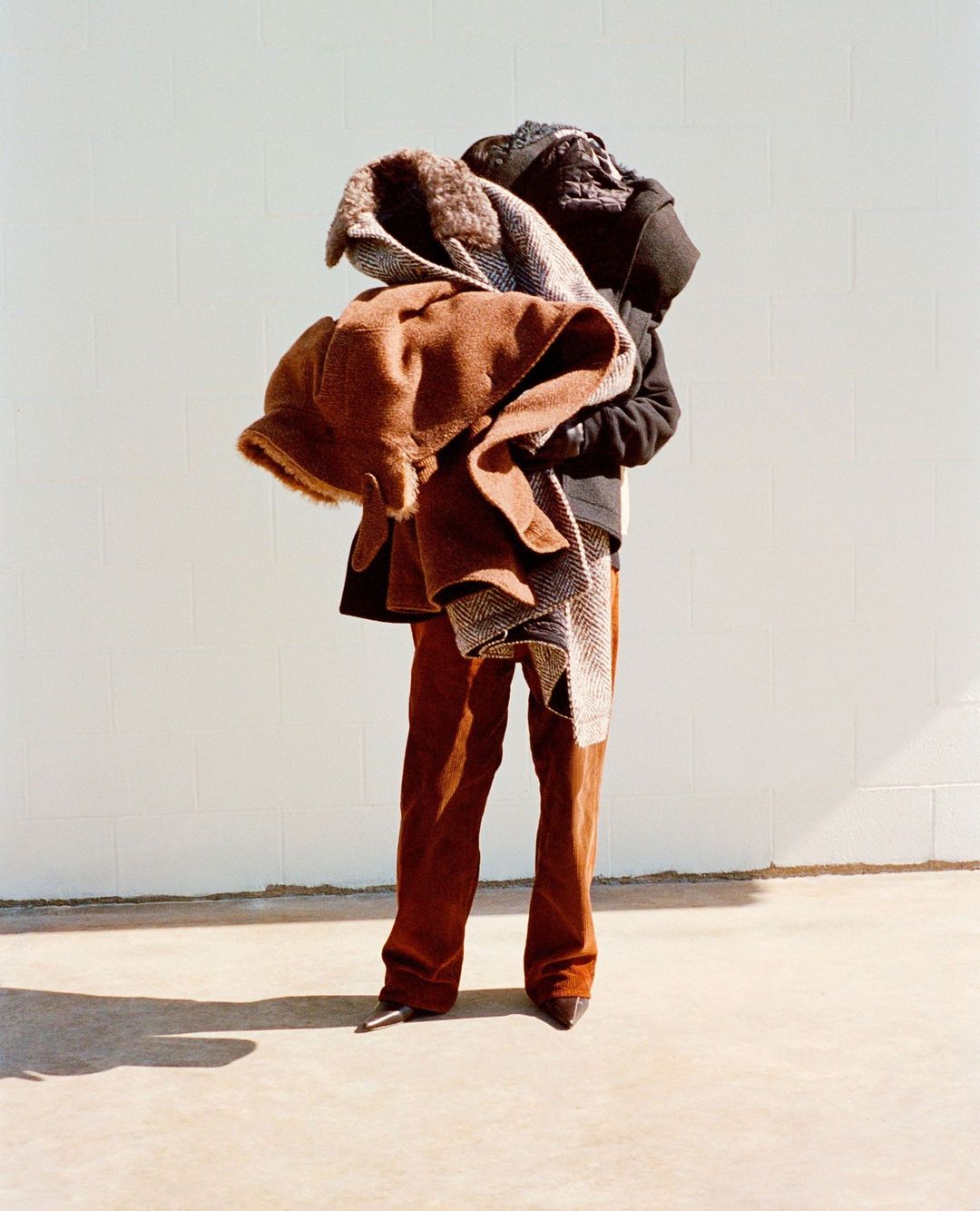 Person stands outdoors holding pile of coats, wearing brown corduroy pants and dark pointy shoes against a light gray wall backdrop.