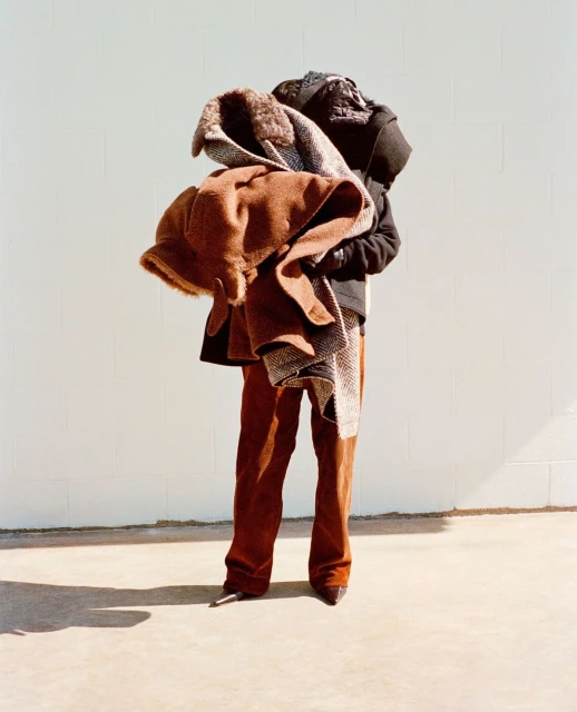 Person stands outdoors holding pile of coats, wearing brown corduroy pants and dark pointy shoes against a light gray wall backdrop.