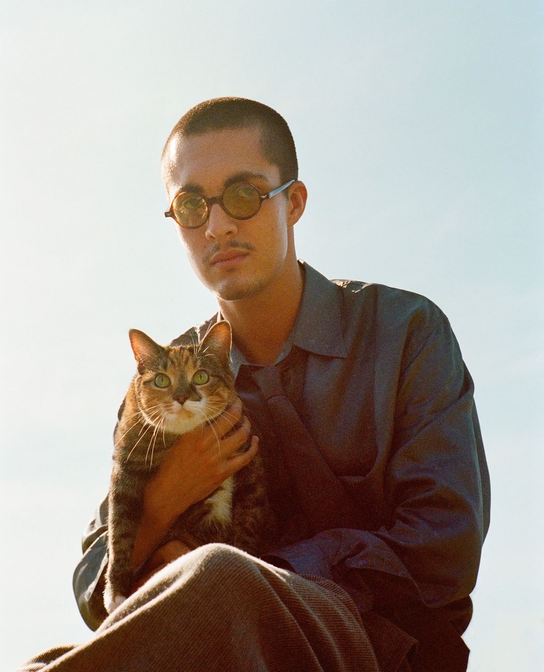A young adult male with buzzcut hair, wearing round sunglasses and a collared shirt, holds a tabby cat in an outdoor setting.
