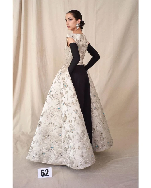 Woman in elegant gown with floral design and black arm gloves posing in a studio, beauty shot with neutral backdrop.