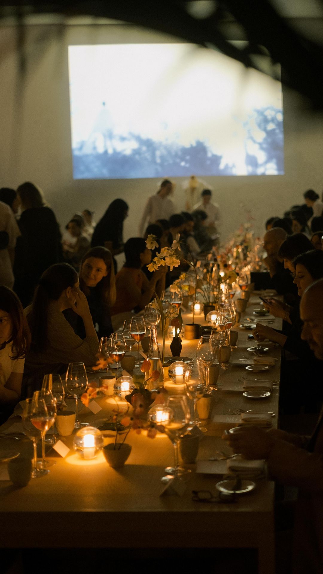 Long dining table with glowing candles and wine glasses at an event with many people seated, a projector screen shows a soft image.