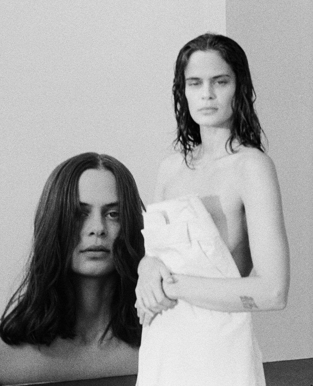 Black and white portrait of two androgynous adults, one woman partially draped in white fabric, posing indoors with a simple wall background.
