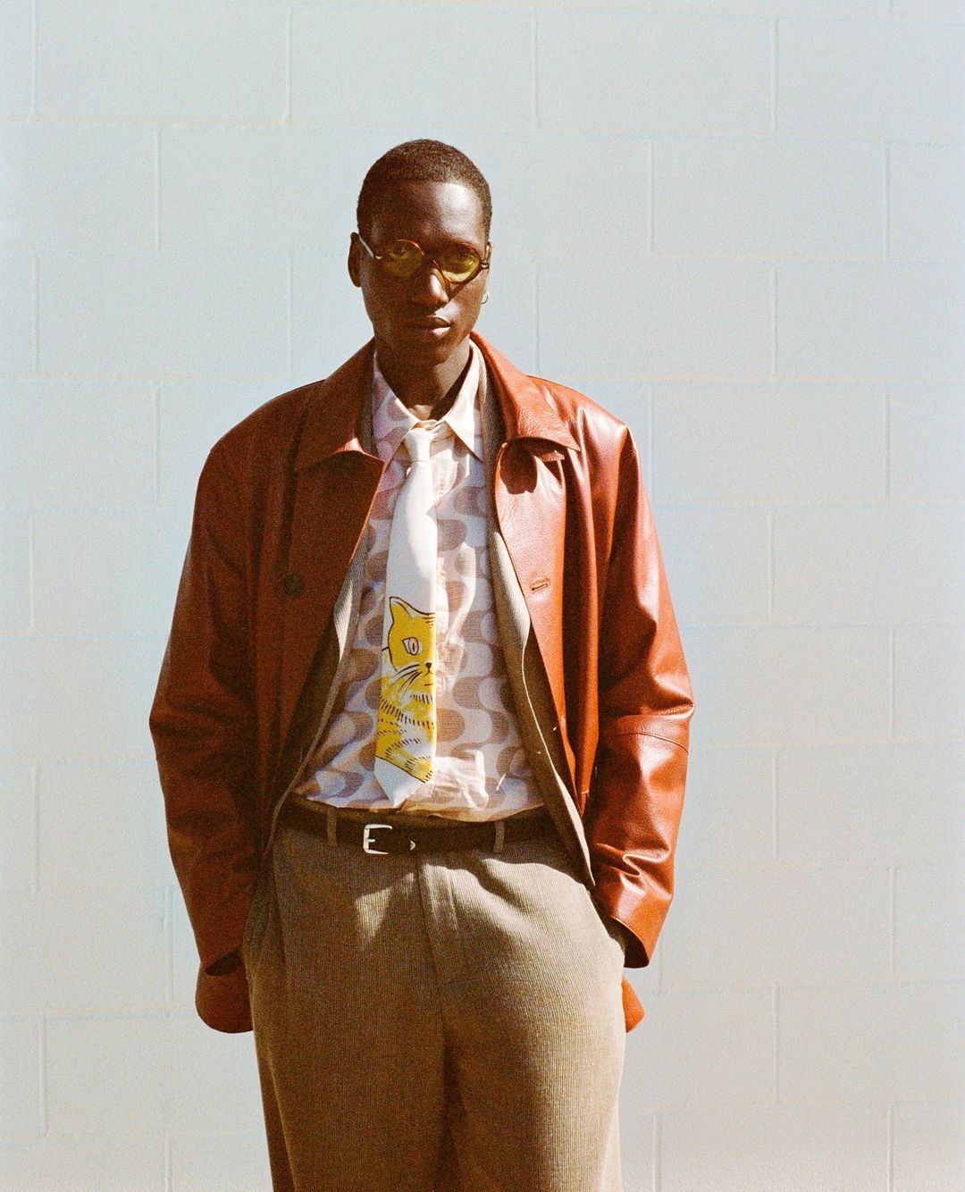 A slender black man in a brown leather jacket and stylish yellow sunglasses stands in front of a white brick wall, modeling a patterned shirt and tie.