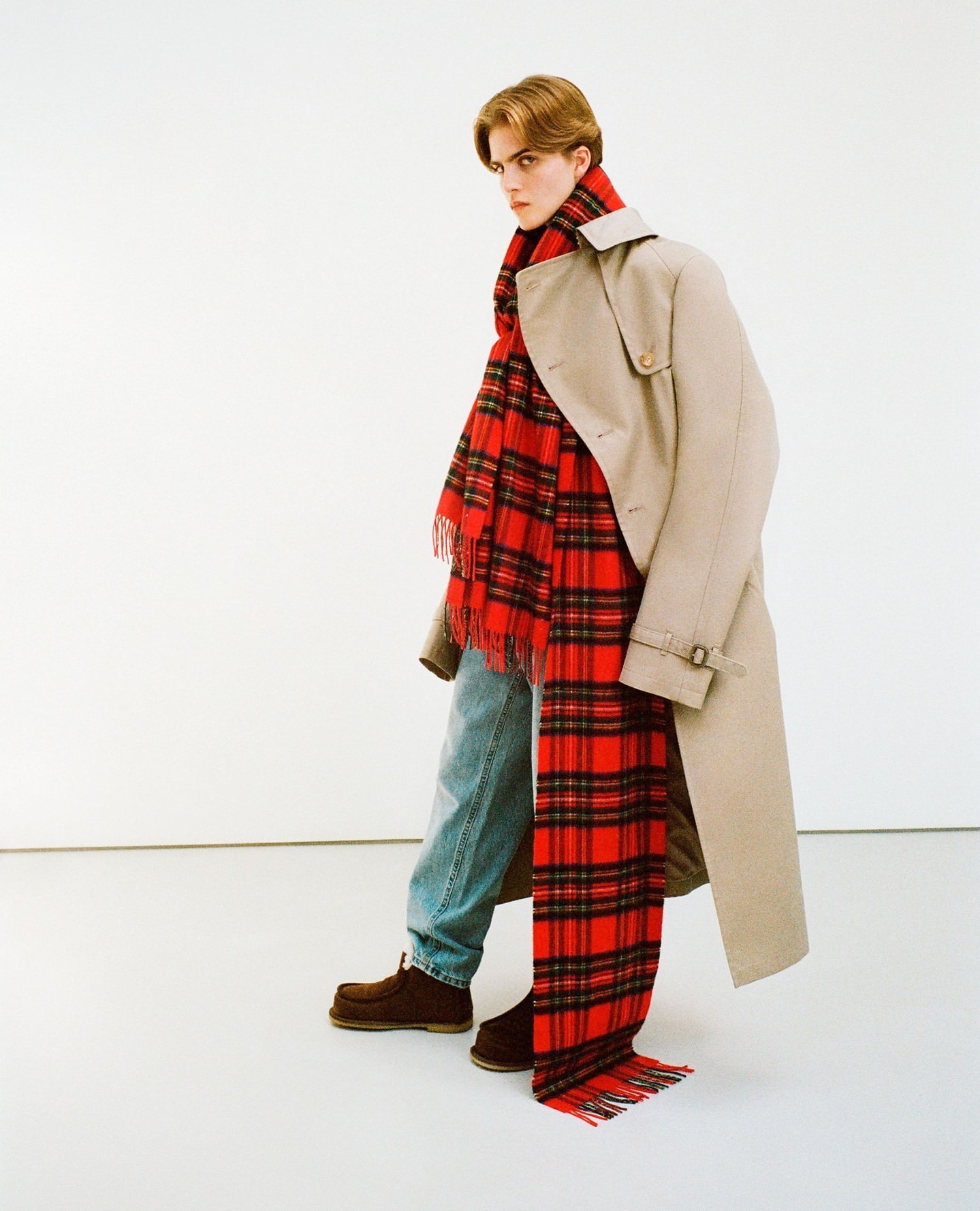 Male model poses in a beige trench coat and red plaid scarf against a white backdrop, showcasing a blend of classic and contemporary style.
