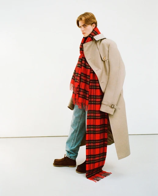 Male model poses in a beige trench coat and red plaid scarf against a white backdrop, showcasing a blend of classic and contemporary style.