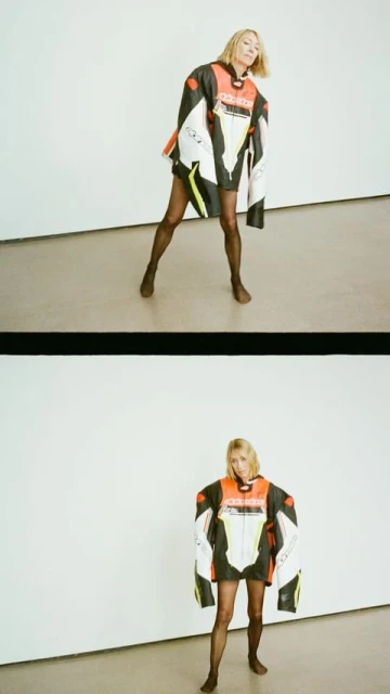 Woman posing in an oversized racing jacket, dark tights, and brown shoes against a white wall and beige floor in a photo studio.
