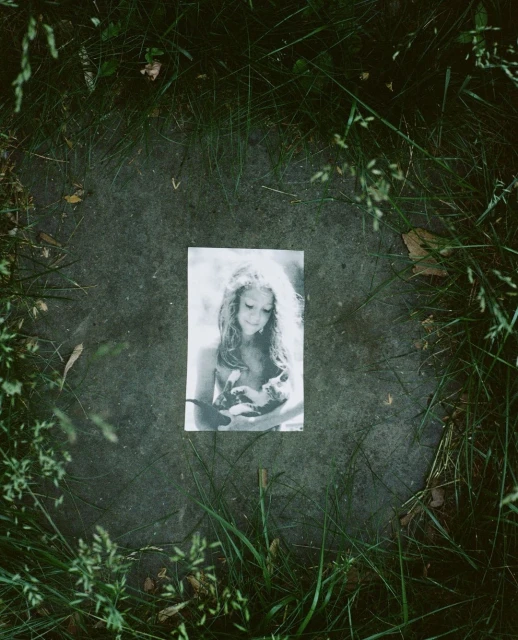 Monochrome photograph of a young girl holding a bird, placed on a stone surface surrounded by green grass, creating a natural, outdoor setting.