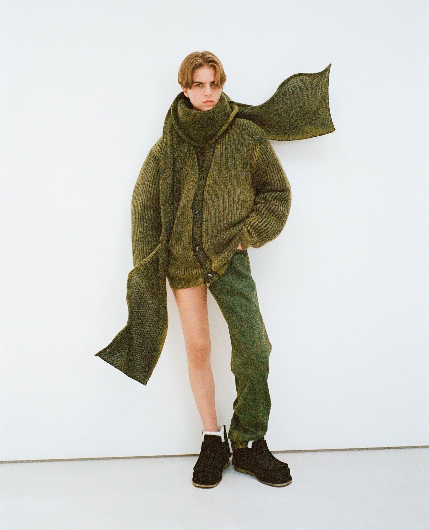 A young adult poses in a green cardigan, scarf, and pants against a white background, showcasing a unique, layered fashion ensemble.