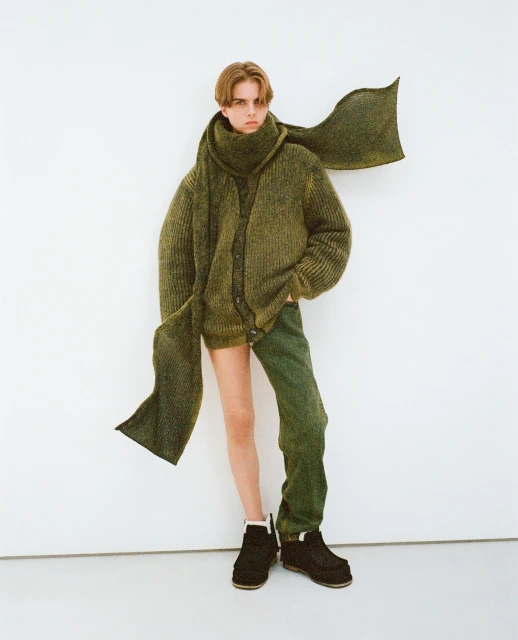A young adult poses in a green cardigan, scarf, and pants against a white background, showcasing a unique, layered fashion ensemble.