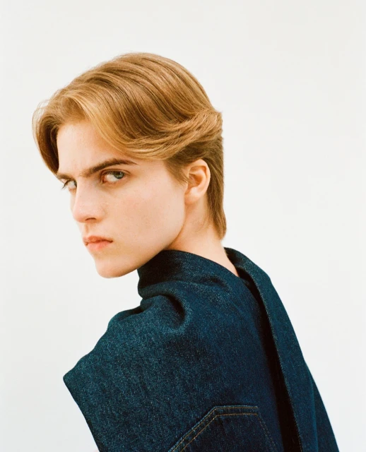Headshot of a young adult with blond hair in a blue denim shirt, posing in front of a white background while looking over shoulder.