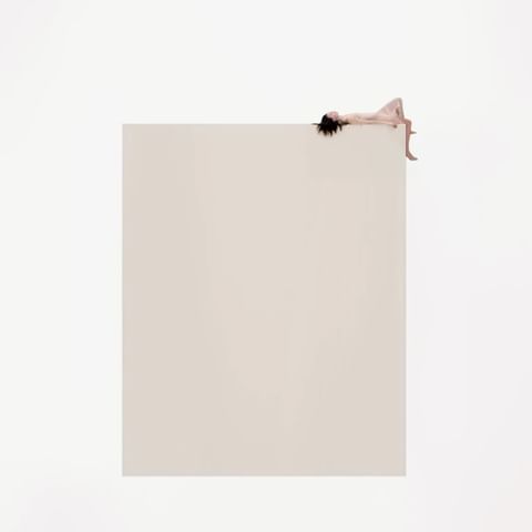 Woman with dark hair reclining at the edge of a large beige surface in a minimalist style against a white background.