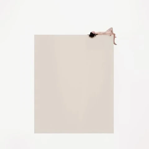 Woman with dark hair reclining at the edge of a large beige surface in a minimalist style against a white background.