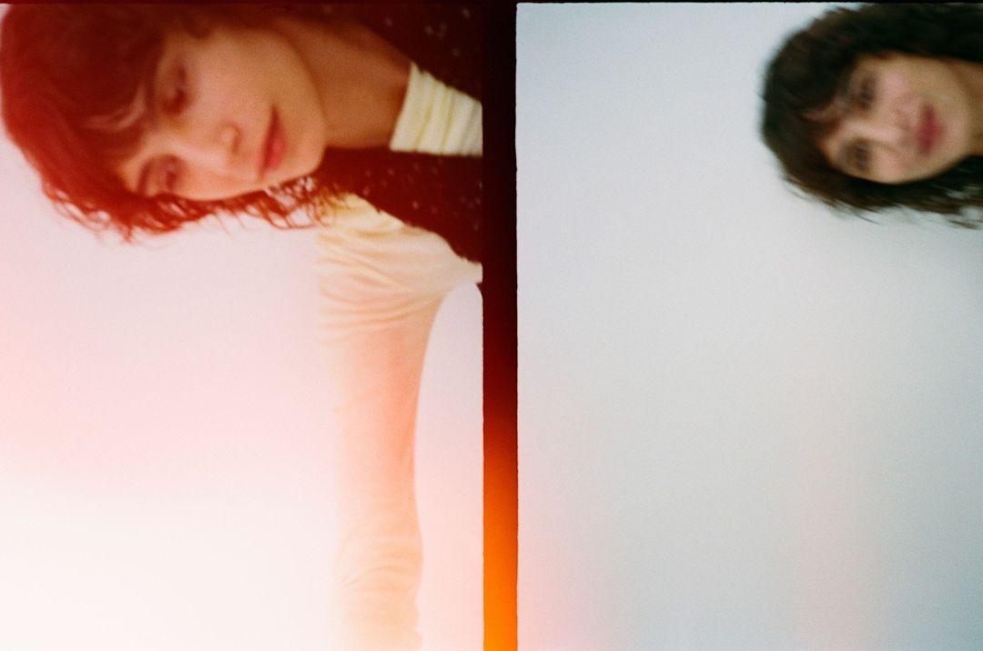 Two women in a portrait photo; one looks down with an orange flare; the other is unfocused in the background on a white surface.