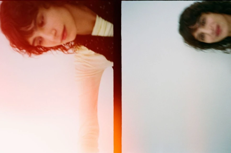 Two women in a portrait photo; one looks down with an orange flare; the other is unfocused in the background on a white surface.