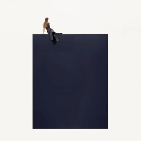 A woman in a black dress sits on the edge of a dark blue block against a white background in a minimalist and abstract composition.