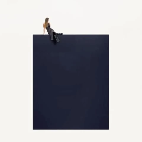 A woman in a black dress sits on the edge of a dark blue block against a white background in a minimalist and abstract composition.