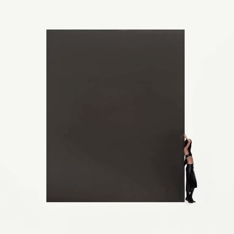 Woman posing in black attire stands behind a large, dark gray rectangular solid against a white background in a studio setting.