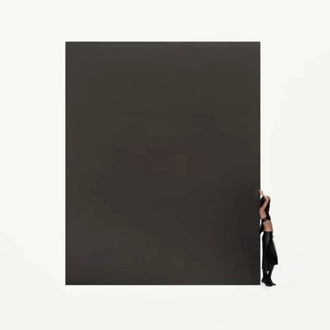 Woman posing in black attire stands behind a large, dark gray rectangular solid against a white background in a studio setting.