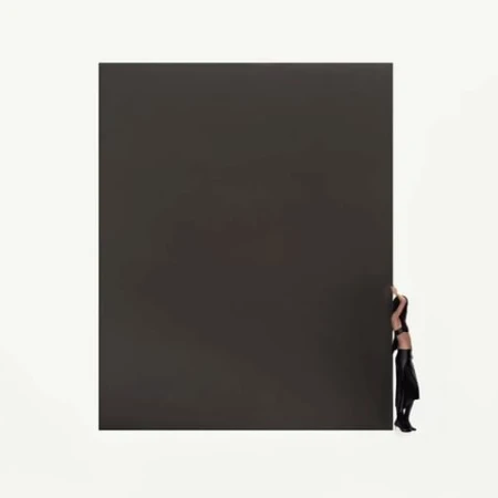 Woman posing in black attire stands behind a large, dark gray rectangular solid against a white background in a studio setting.