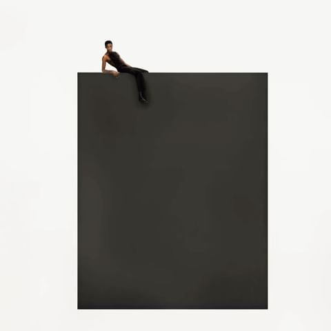Adult male reclines on the top edge of a dark grey cube against a stark white backdrop in a minimalist studio-style composition.