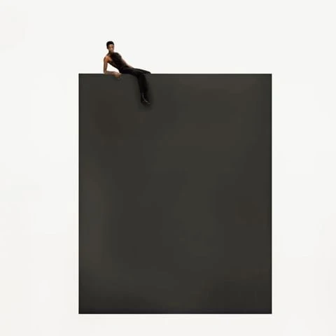 Adult male reclines on the top edge of a dark grey cube against a stark white backdrop in a minimalist studio-style composition.