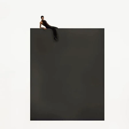 Adult male reclines on the top edge of a dark grey cube against a stark white backdrop in a minimalist studio-style composition.
