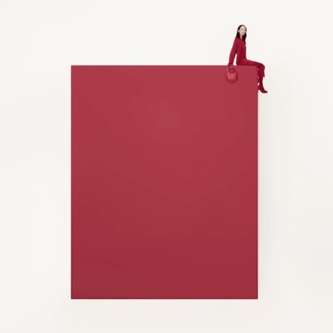 A woman in a red jumpsuit sits on the corner of a large red square against a white backdrop, holding a matching red purse.