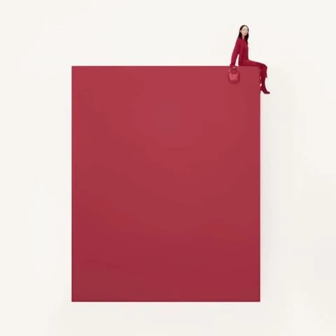 A woman in a red jumpsuit sits on the corner of a large red square against a white backdrop, holding a matching red purse.