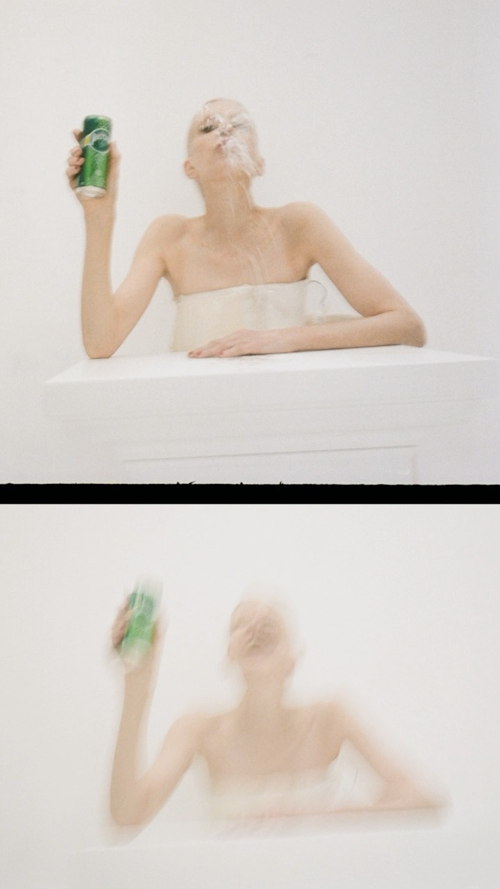 A woman with short hair sits at a white table, holding a green can while smoke comes from her mouth in a bright, clean environment.