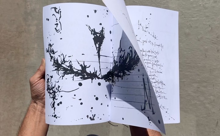 Open book features abstract black ink blot art on one page and handwritten text on the other, held by adult human hands.