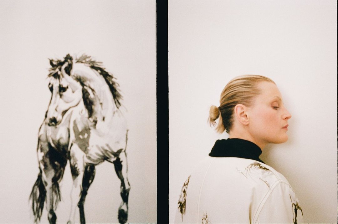 Adult female poses in a photo studio with a black-and-white horse painting. The woman wears a printed jacket and faces the wall.