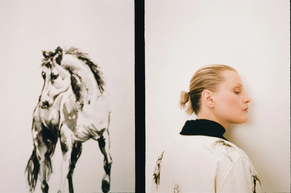 Adult female poses in a photo studio with a black-and-white horse painting. The woman wears a printed jacket and faces the wall.