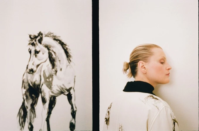 Adult female poses in a photo studio with a black-and-white horse painting. The woman wears a printed jacket and faces the wall.
