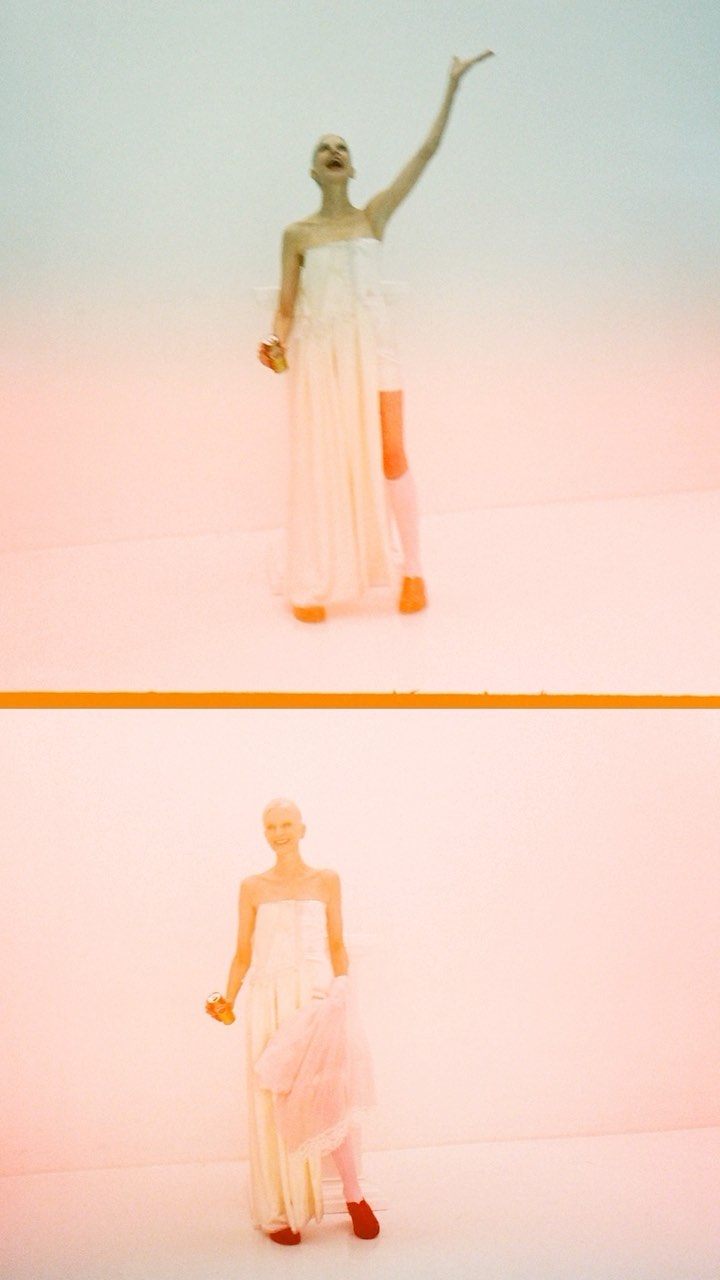 Two fashion models in white gowns, one arm raised, stand against a white background with a thin orange line dividing them.