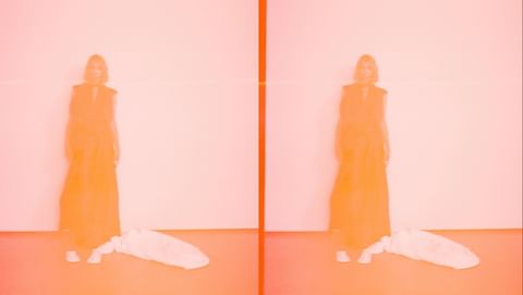 A woman in a sleeveless dress stands in a studio with a bag on the floor, bathed in orange light and captured with a blurry aesthetic.