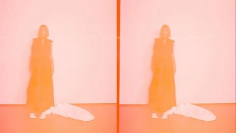 A woman in a sleeveless dress stands in a studio with a bag on the floor, bathed in orange light and captured with a blurry aesthetic.