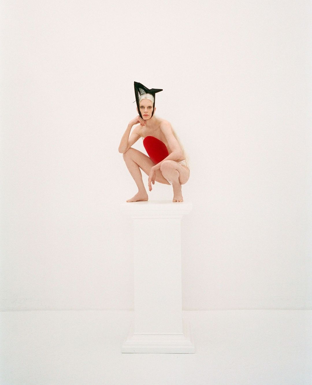 Elegant woman with pale skin and dramatic black headpiece in a crouching pose atop a white column in a brightly lit white photo studio.