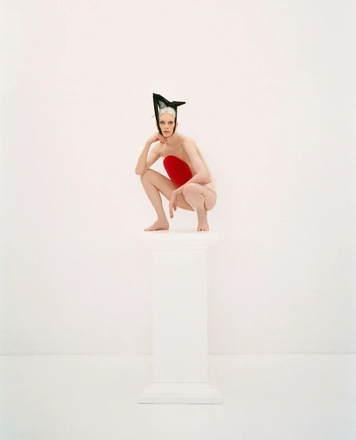 Elegant woman with pale skin and dramatic black headpiece in a crouching pose atop a white column in a brightly lit white photo studio.