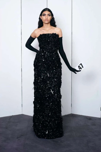 A woman stands in a photo studio wearing a long, strapless black gown and long black gloves, holding a card with the number 47.