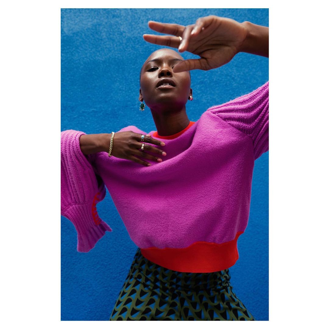 Headshot of a fashionable Black woman in a pink sweater, geometric-patterned skirt, and jewelry, posing against a bright blue background.