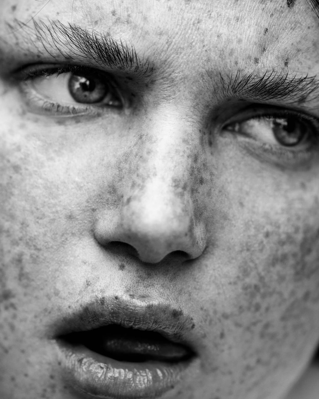 Close-up black and white portrait of a freckled adult female's face with her tongue slightly extended, captured in a studio environment.