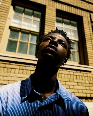 Low-angle shot of a young Black man wearing a blue striped button-down shirt outdoors against a brick building.