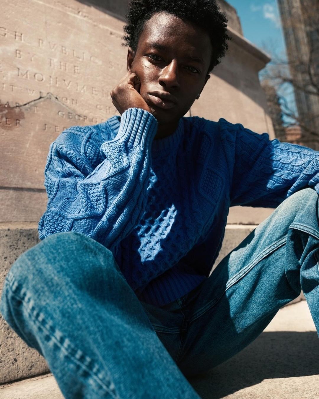 Young man poses outdoors in a blue knit sweater and jeans, sitting against a stone structure in sunlight, creating a fashion editorial image.