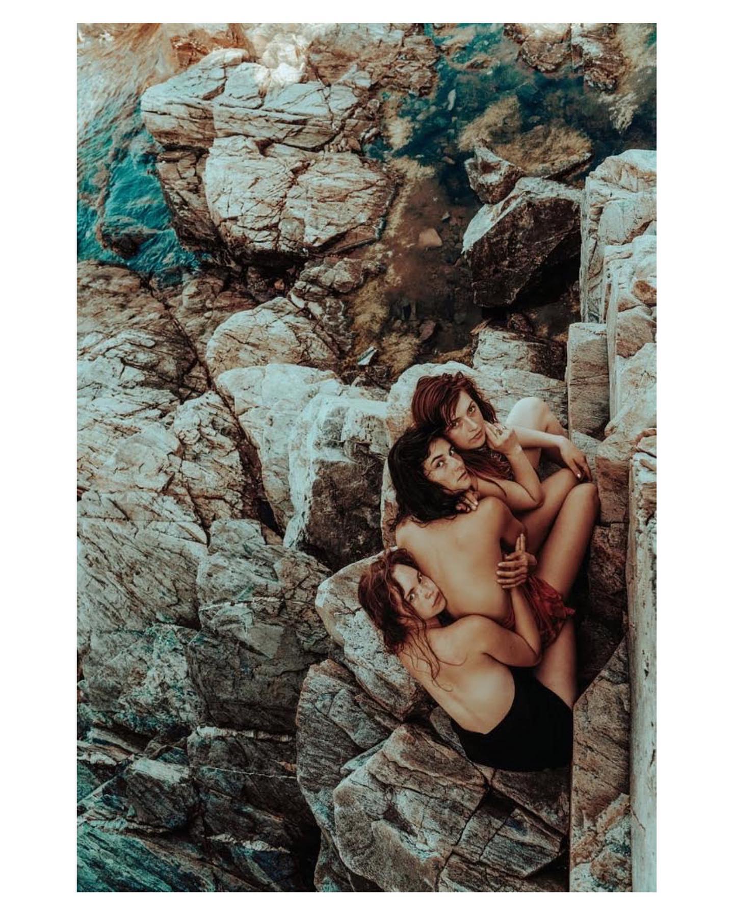 Three women pose nude together on a rocky beach with turquoise water, creating a natural fashion image. The group hugs close together.