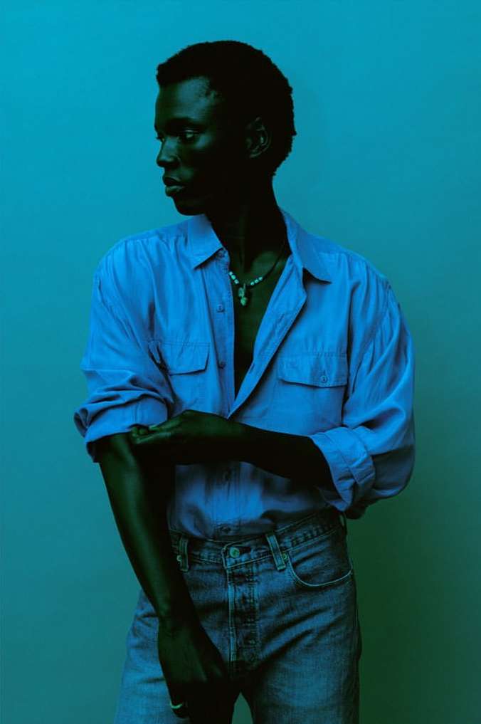 Portrait of a dark-skinned man in a blue shirt and jeans, rolling up his sleeve against a gradient blue background. He wears a beaded necklace.