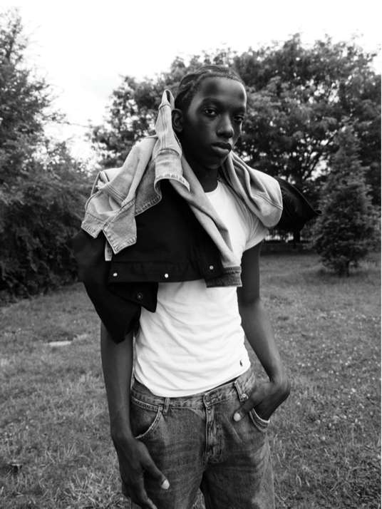 Portrait of a young African American man in casual clothing outdoors, wearing a white t-shirt and jeans with a denim jacket on his shoulder.