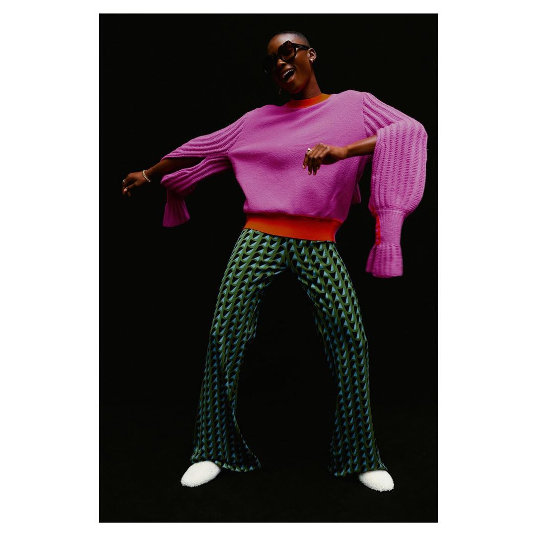A Black fashion model strikes a dynamic pose in a magenta sweater and patterned pants against a black background in a studio shot.
